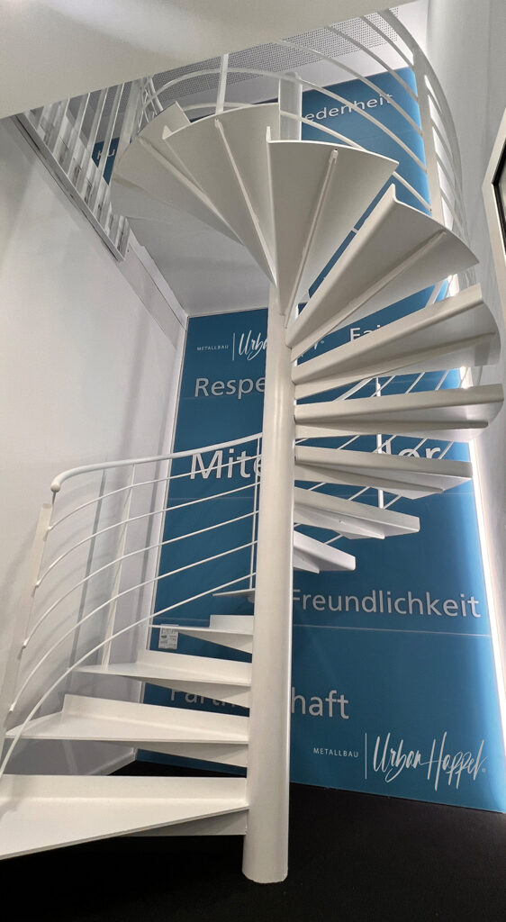 Wendeltreppe Volksbank Laichingen Weiße Wendeltreppe aus Stahl, vor Ort geschweißt, im Innenbereich der Volksbank Laichingen. Die Treppe verbindet das erste und zweite Obergeschoss und wurde passgenau in eine begrenzte Raumhöhe integriert. Im Hintergrund befinden sich Firmenwerte