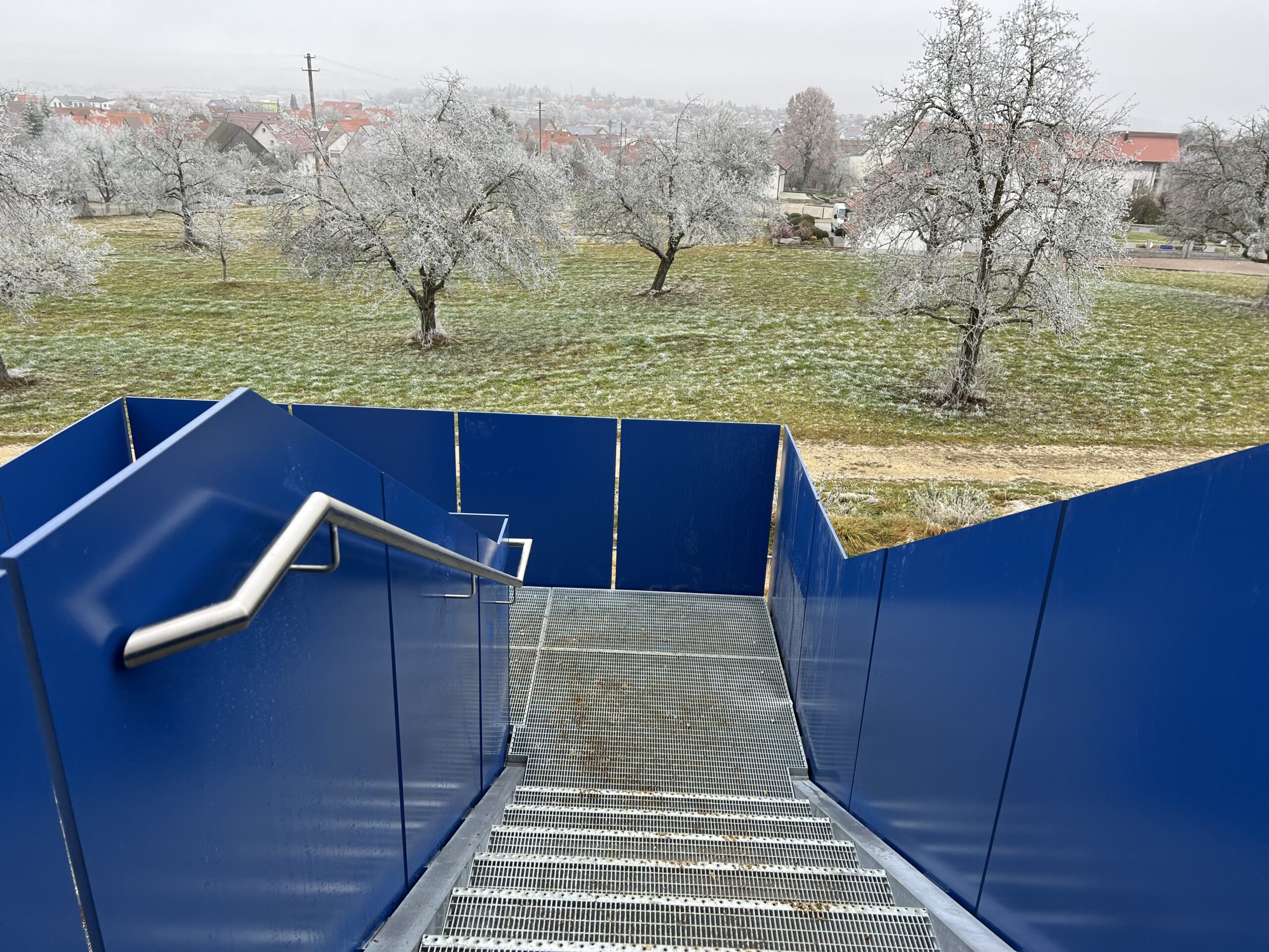 Eine Treppe mit Gitterroststufen und ein Edelstahl Handlauf an einem Blauen Vollblechgeländer