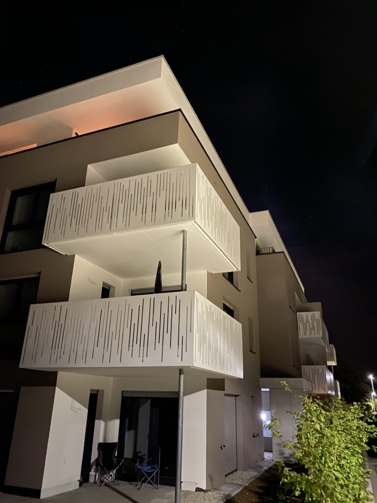 Nachtbild Hintenburgstraße Nachtaufnahme eines Mehrfamilienhauses mit individuell gefrästen Balkonverkleidungen. Die Geländerelemente bestehen aus einzeln gestalteten Aluminiumplatten – jede Ausfräsung ist einzigartig und nicht wiederholt. Die indirekte Beleuchtung betont das besondere Design.