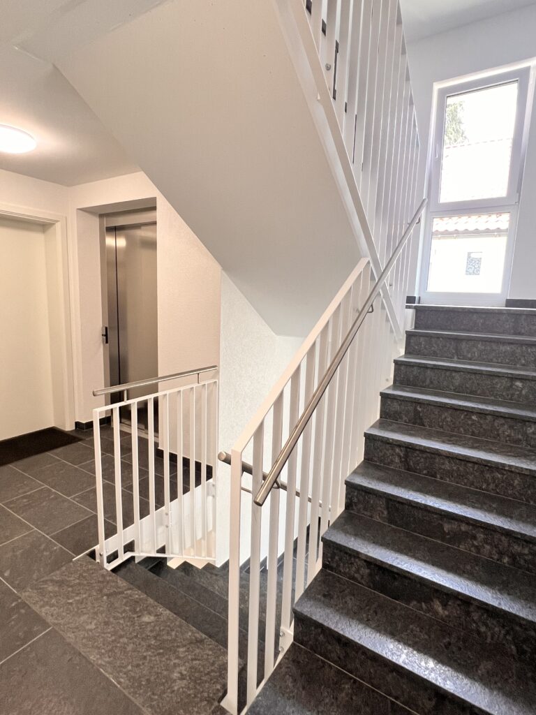 Karl Straße Innengeländer Innenraumaufnahme eines modernen Treppenhauses mit individuell gefertigtem Stahlgeländer. Besonderheit: Die vertikalen Stäbe der Geländerelemente sind im Treppenauge übereinander statt nebeneinander montiert. Die Treppe führt entlang dunkler Naturstein-Stufen mit Edelstahlhandlauf.