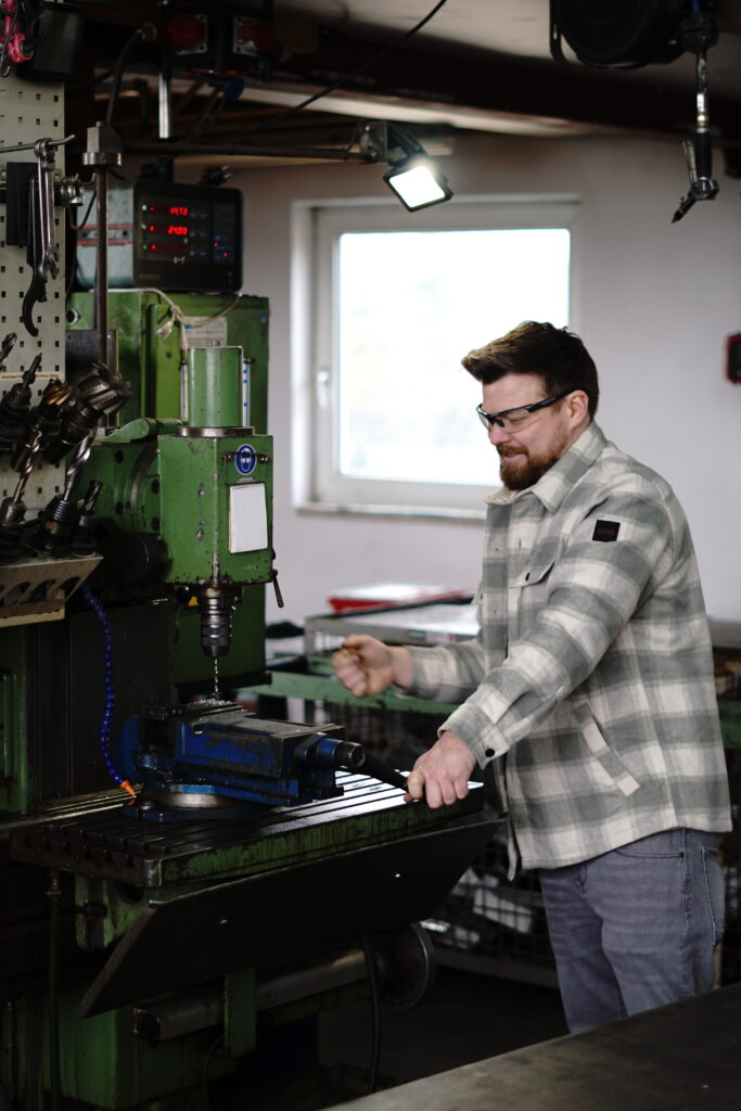 Silvester Happel arbeitet in seiner Werkstatt in Heroldstatt an einer grünen Wemas-Fräsmaschine aus dem Jahr 1972. Mit Schutzbrille und karierter Jacke steht er konzentriert an der Maschine, während Metall bearbeitet wird. Die Werkstatt vermittelt handwerkliche Authentizität und Erfahrung.