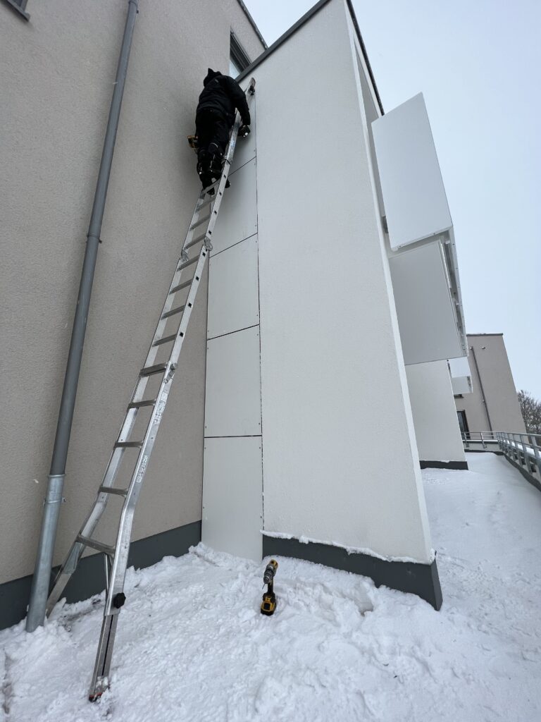 Montage von Trennwänden aus HPL-Platten an einer Außenfassade im Winter. Ein Monteur arbeitet auf einer Leiter in Schneelandschaft, Werkzeug und Material sind im unteren Bildbereich sichtbar. Die Trennwände werden bündig an die Gebäudestruktur angepasst.
