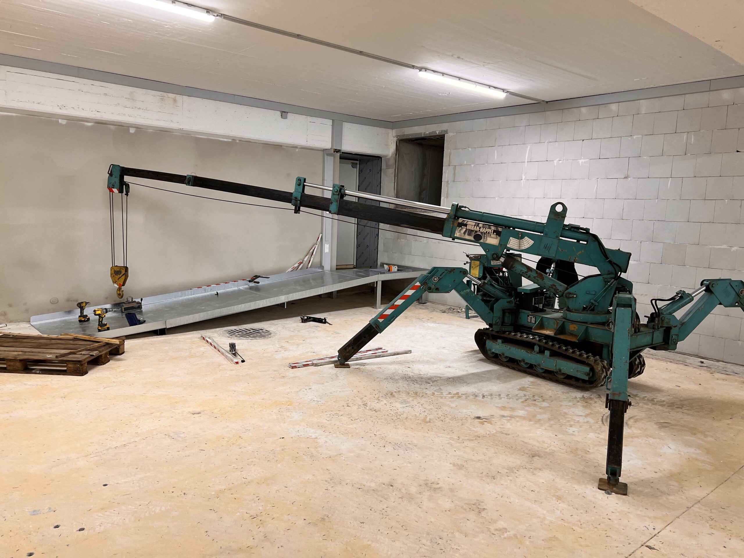 Montage einer barrierefreien Rampe aus verzinktem Blech in einer Tiefgarage. Die Rampe wird mithilfe eines kompakten Raupenkrans mit Teleskoparm positioniert. Auf dem Boden liegen Werkzeuge und Baumaterialien.